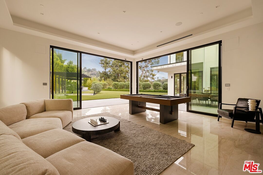 This spacious, open-concept living area features a large, light-colored sectional sofa arranged around a circular coffee table with a chess set. A pool table sits in the center of the room, which is framed by expansive floor-to-ceiling glass sliding doors that provide a seamless transition to the outdoor lawn. The room is finished with polished light-colored flooring and a neutral color palette, creating a bright and airy atmosphere suitable for entertaining.