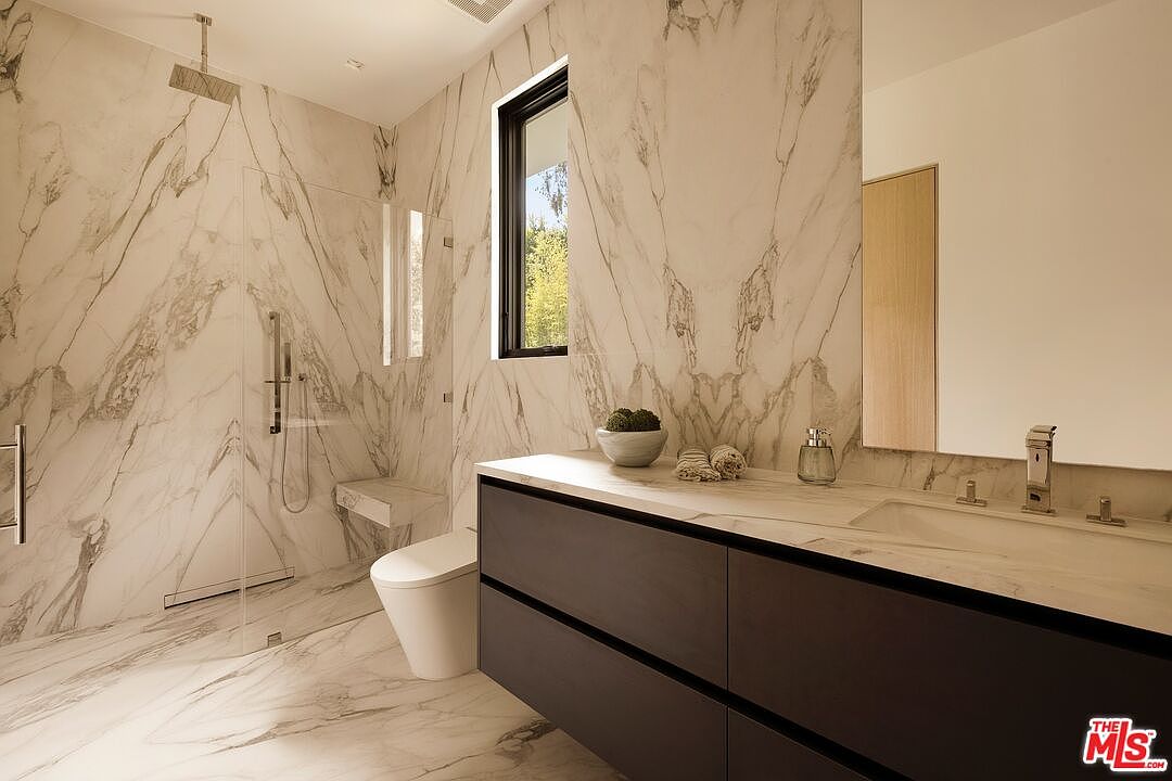 This modern primary bathroom features extensive use of light-toned stone with dramatic veining on the walls and floors. The space includes a floating vanity with dark cabinetry, a large wall-mounted mirror, and a walk-in shower enclosure with a built-in bench. A tall, narrow window provides natural light, creating a bright and sophisticated atmosphere.