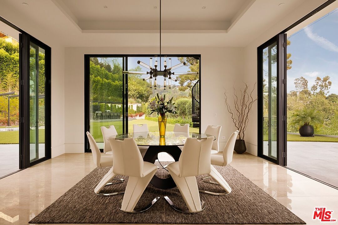 This elegant dining room features a circular glass-topped table surrounded by six modern white chairs, all set upon a textured area rug. The space is framed by expansive glass doors that provide a seamless transition to the outdoor landscape, while a contemporary sputnik-style chandelier serves as a striking focal point above. The room's neutral color palette and clean lines create a sophisticated and airy atmosphere perfect for formal entertaining.