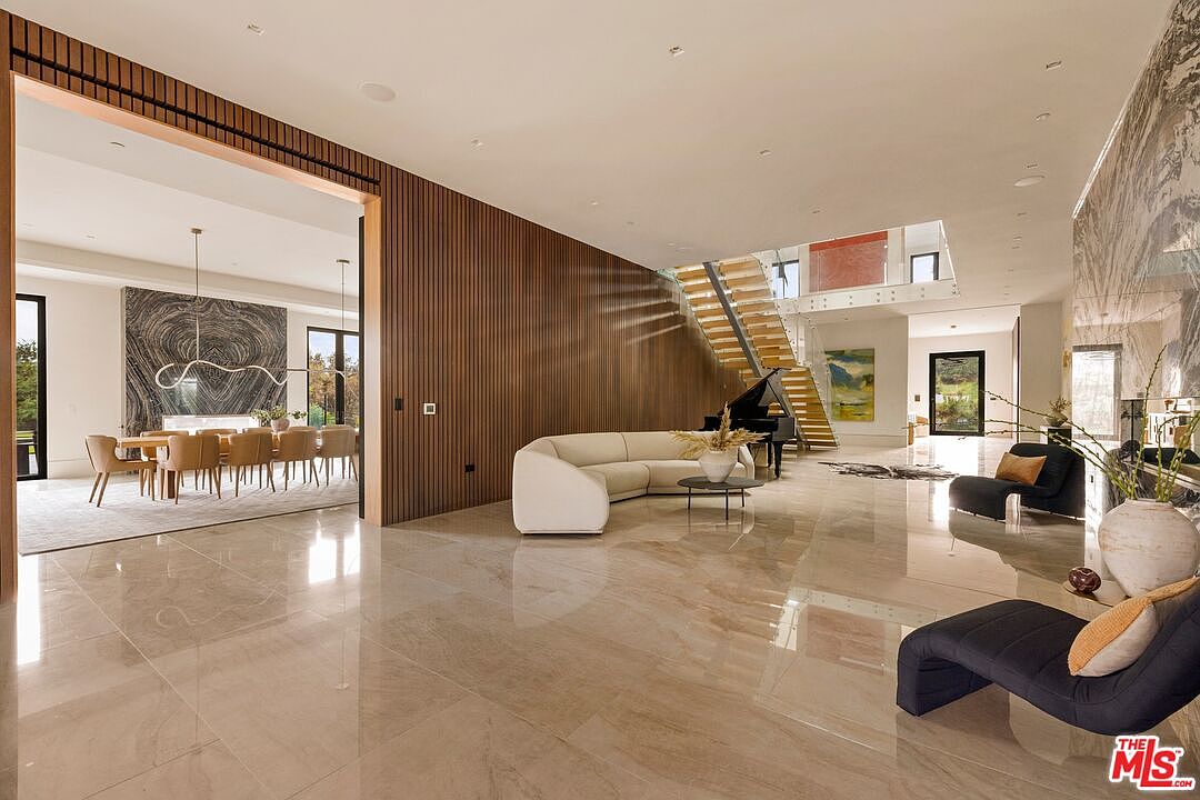 This expansive, open-concept living area features polished stone flooring and a striking wood-slat accent wall that separates the main space from the adjacent dining room. A curved, cream-colored sofa sits centrally before a grand piano and a floating staircase, creating a sophisticated and airy atmosphere. The perspective captures the seamless flow between the formal living area and the dining room, which is anchored by a dramatic, dark-veined stone wall feature.