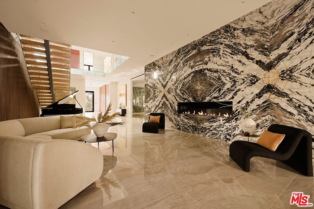 This expansive living area features a striking, book-matched stone wall with a linear fireplace insert as its central focal point. The room is furnished with a curved, light-toned sofa and two modern black lounge chairs, all set upon a polished, reflective stone floor. A floating staircase with light wood treads and glass railings adds a contemporary architectural element to the open-concept space.