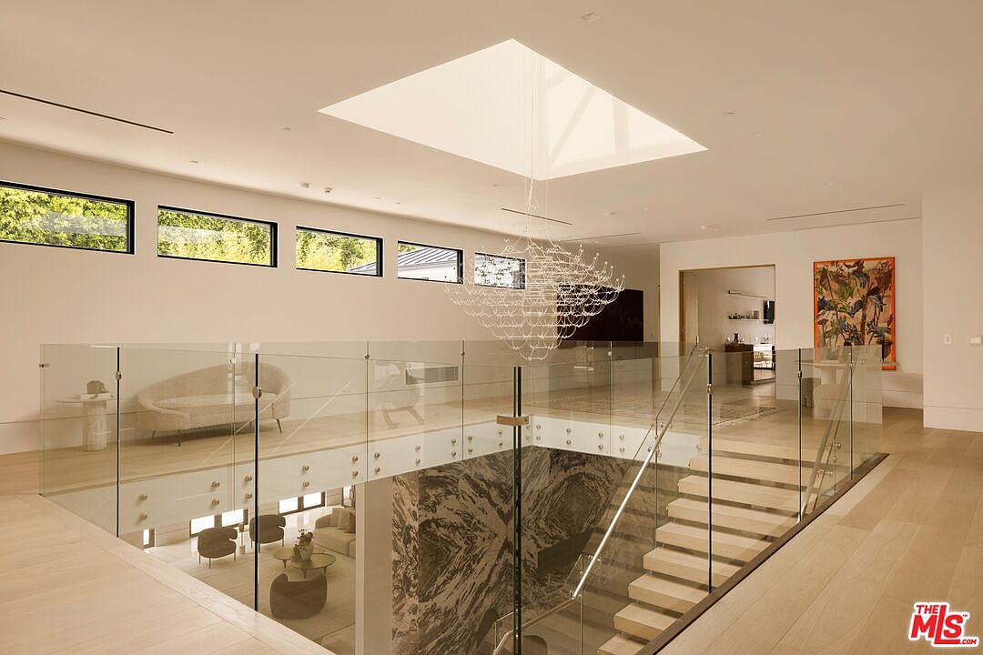 This modern, open-concept landing area features a striking glass-railed staircase that overlooks a lower level with a dramatic, dark-veined stone feature wall. A large, sculptural chandelier hangs beneath a central skylight, casting soft light over the light-toned wood flooring. The space is minimalist and airy, with a series of horizontal windows providing natural light and a view of the outdoors.