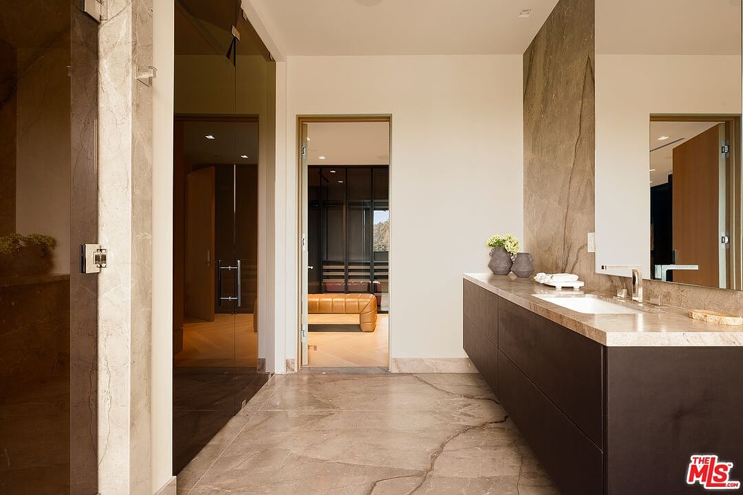 This modern primary bathroom features a sleek vanity with a dark base and a light stone countertop, complemented by a large mirror and a tall stone-clad wall section. The space is finished with large-format stone flooring and leads into a walk-in closet area visible through an open doorway. The perspective captures the clean lines and high-end finishes of the room, emphasizing a sophisticated and minimalist aesthetic.