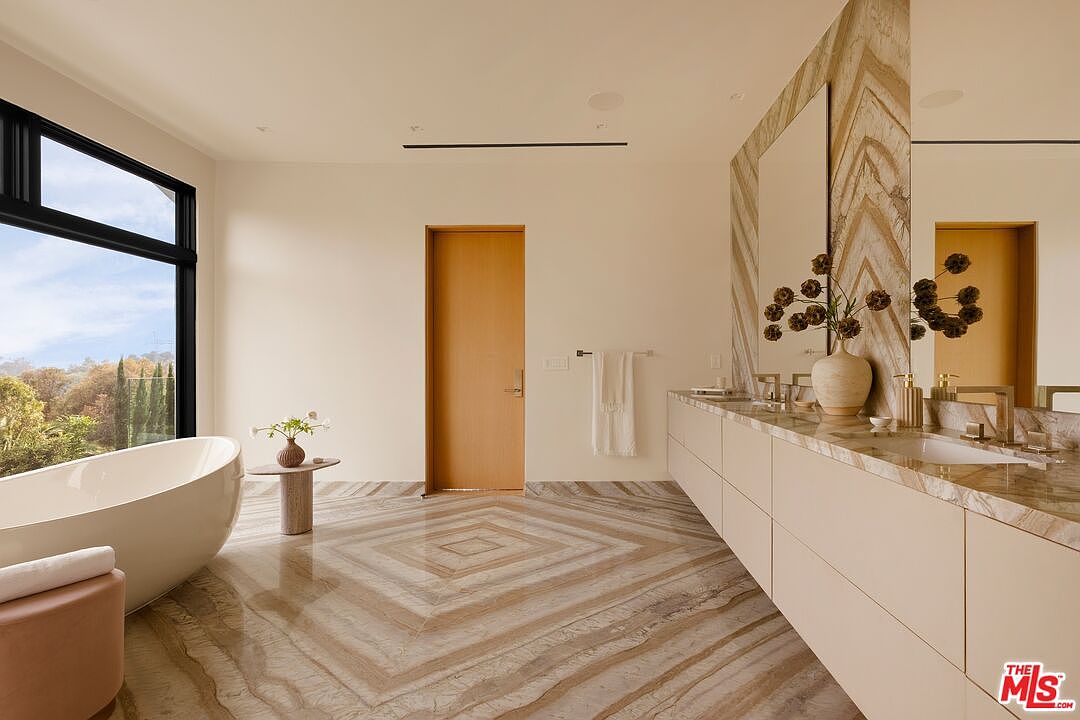 This luxurious primary bathroom features a striking, patterned stone floor that extends up the wall behind the vanity, creating a cohesive and high-end aesthetic. A sleek, freestanding soaking tub is positioned near large windows that offer expansive views of the surrounding landscape, while a long, light-toned vanity provides ample counter space. The room is bathed in natural light, emphasizing the clean lines and sophisticated, minimalist design.