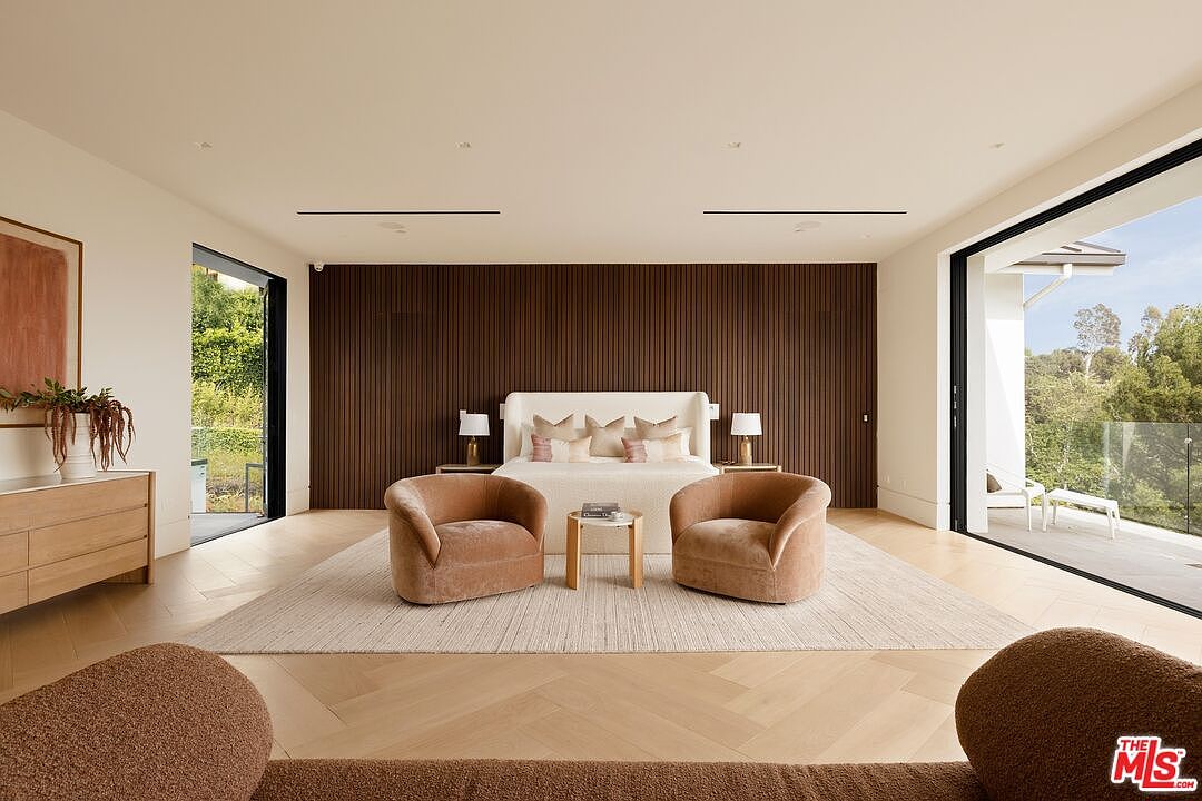 This spacious primary bedroom features a prominent dark wood-slatted accent wall behind a neutral-toned bed. The room is framed by large glass sliding doors on either side that open to outdoor views, and it includes a pair of plush, curved armchairs arranged around a small side table. The light wood flooring and minimalist decor create a serene, high-end atmosphere.