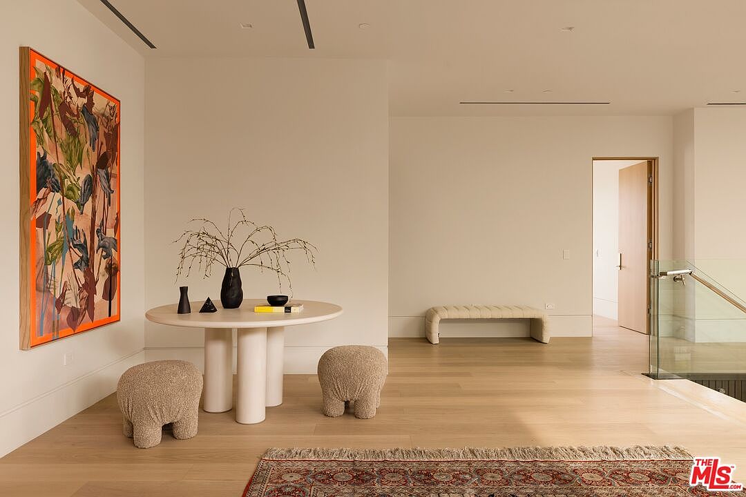 This bright, minimalist entryway or landing area features light wood flooring and a neutral color palette. A round pedestal table sits in the center, flanked by two textured, rounded stools, while a large, vibrant abstract painting adds a bold focal point to the wall. To the right, a glass-railed staircase leads to another level, and a doorway opens into an adjacent room, creating an open and airy transition space.