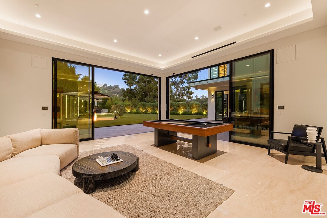 This spacious, modern living area features a large, light-colored sectional sofa and a dark wood coffee table topped with a chess set. A professional-grade pool table serves as a central focal point, while expansive floor-to-ceiling glass sliding doors provide a seamless transition to the outdoor lawn. The room is finished with polished stone flooring and recessed lighting, creating a bright and sophisticated atmosphere.