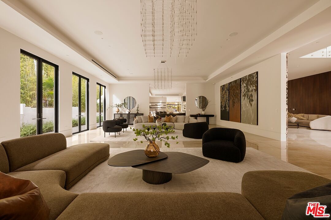 This expansive, open-concept living room features a sophisticated neutral palette with plush, curved seating arrangements centered around a layered, organic-shaped coffee table. Large windows along the left wall provide abundant natural light and views of the exterior greenery, while a striking tiered chandelier serves as a focal point above the seating area. The space is elegantly decorated with large-scale abstract art and minimalist decor, creating a luxurious and airy atmosphere.