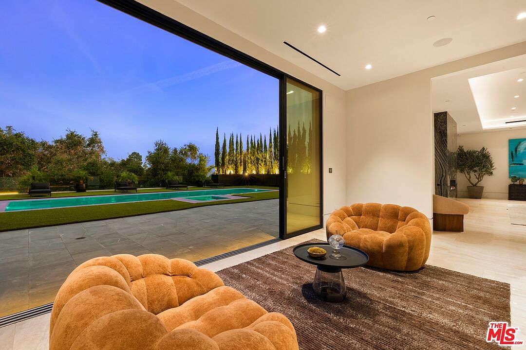 This contemporary living space features two plush, rounded armchairs arranged around a dark circular coffee table on a textured area rug. A massive wall of glass slides open to reveal a seamless transition to a stone patio and a long, rectangular swimming pool set against a backdrop of illuminated cypress trees. The interior is characterized by neutral tones, recessed lighting, and a glimpse into an adjacent hallway featuring a tall, dark stone-clad feature wall.