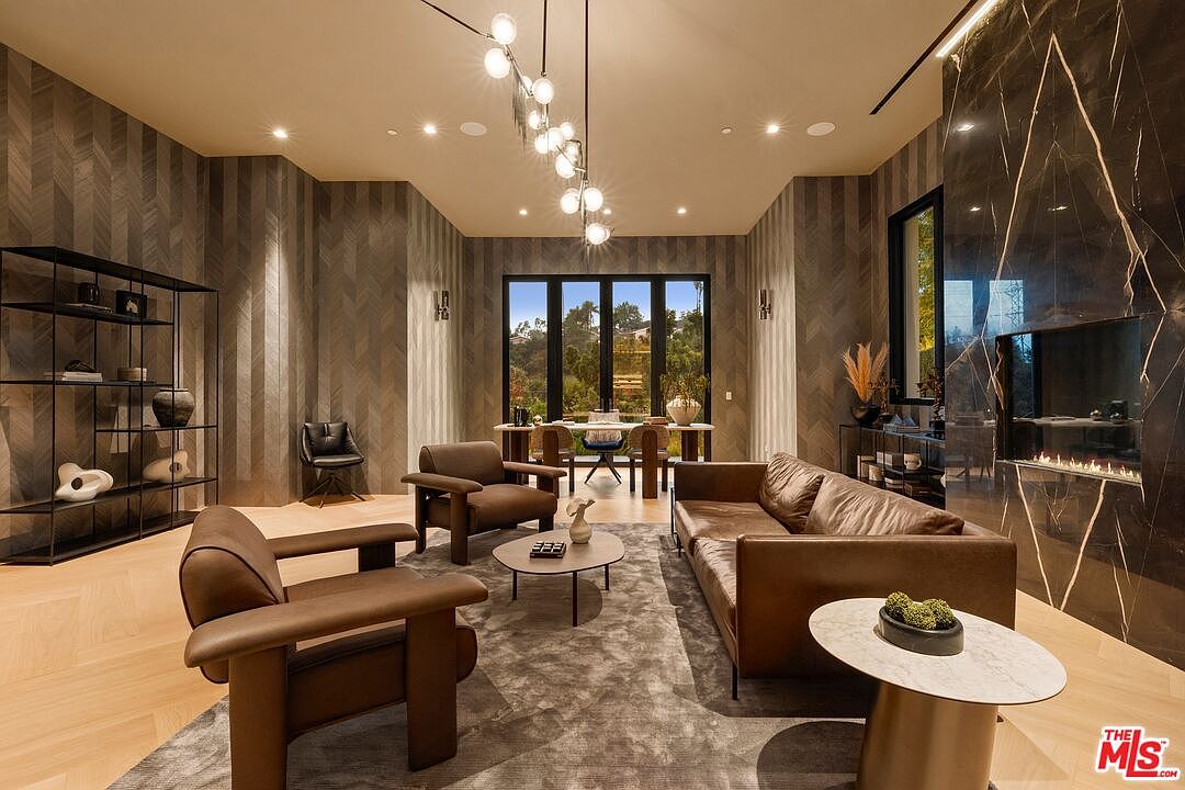 This sophisticated living room features a warm, neutral color palette with textured chevron-patterned wallpaper and light wood flooring. The space is anchored by a leather sofa and matching armchairs arranged around a central coffee table, all set against a striking dark stone-clad chimney breast. A modern, multi-pendant light fixture hangs above the seating area, while large glass doors at the rear provide a view of the outdoor landscape.