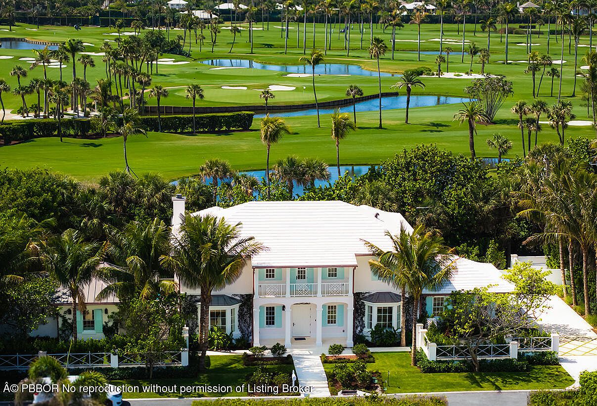 This aerial view showcases a luxurious two-story home with a white roof and light blue shutters, nestled amidst lush greenery and palm trees. The property overlooks a meticulously maintained golf course with water features, creating a serene and exclusive atmosphere. A paved driveway leads to the house, enhancing its curb appeal.