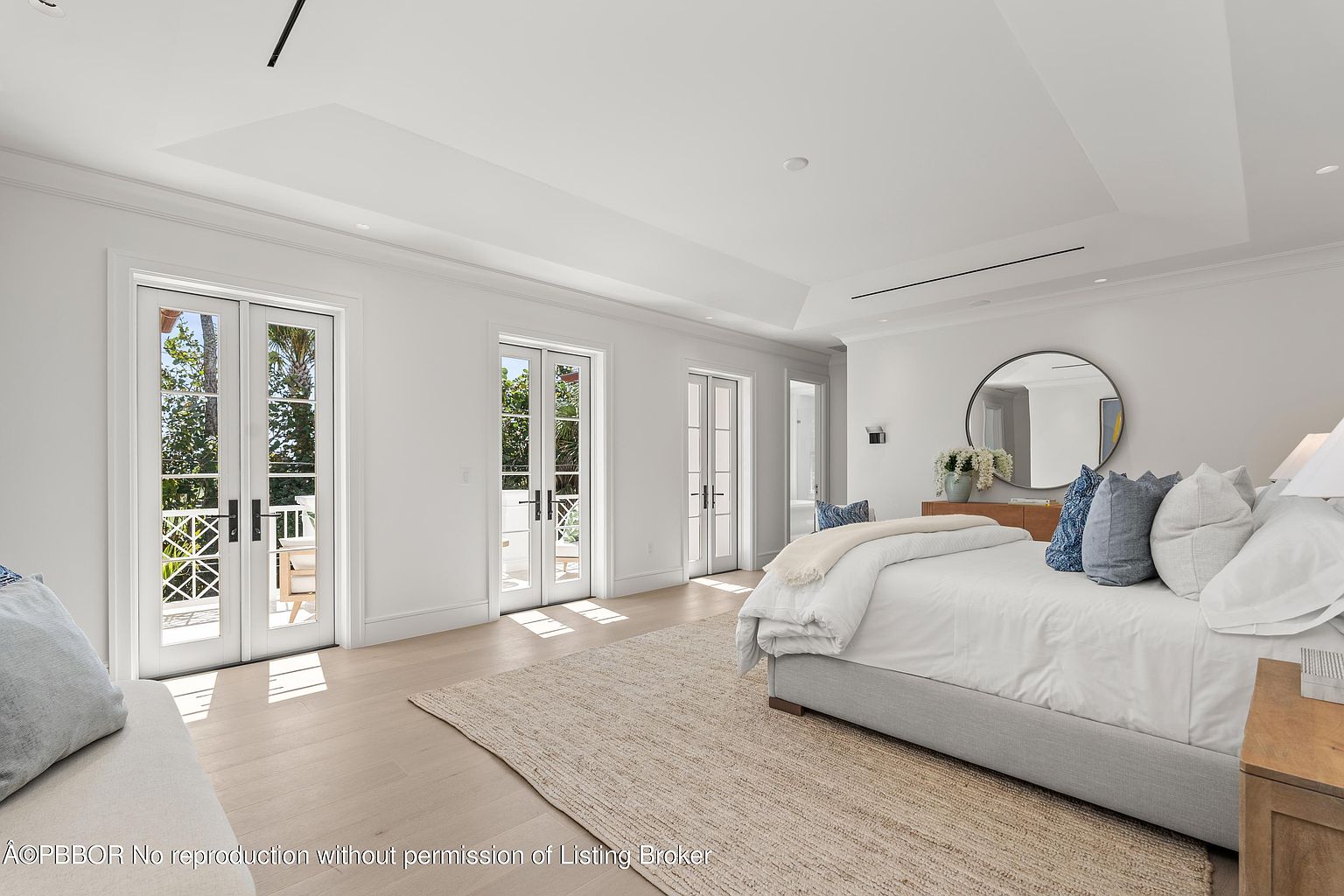 This is a bright and spacious primary bedroom featuring white walls, light wood flooring, and multiple sets of French doors leading to outdoor spaces. The room is furnished with a large bed with white linens and blue pillows, a round mirror above a wooden dresser, and a neutral-toned area rug. The overall aesthetic is clean, modern, and luxurious, emphasizing natural light and a serene atmosphere.