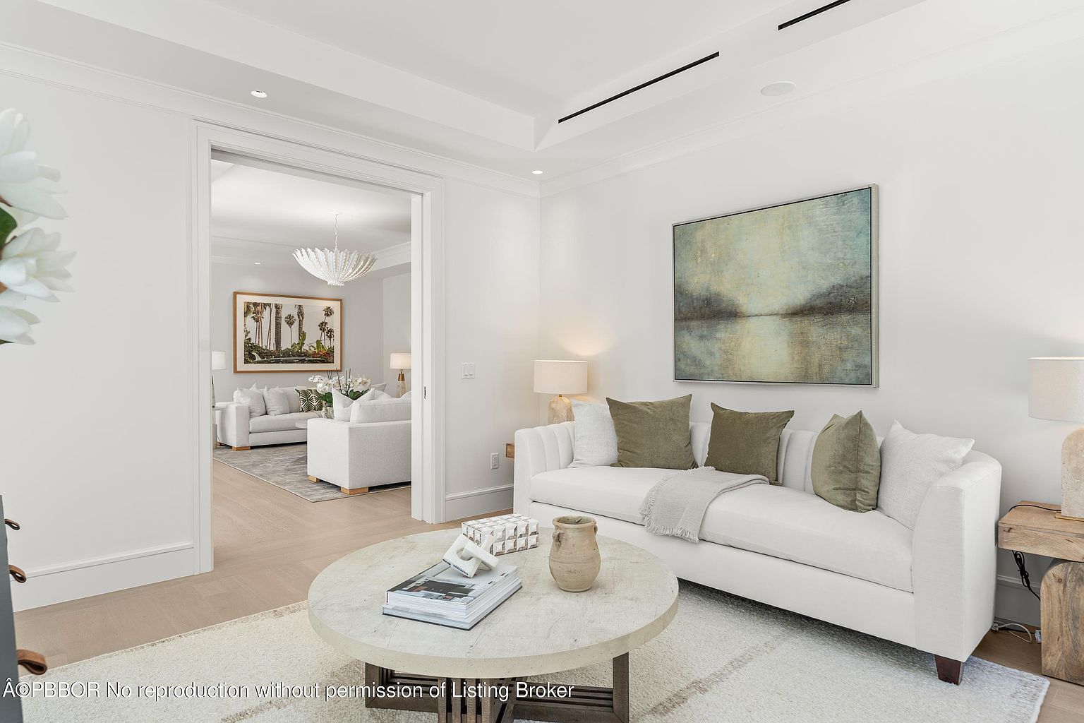 This is an interior shot of a living room featuring a white sofa with olive green pillows, a round marble coffee table with books and decorative items, and an abstract painting on the wall. The room is bright and airy with white walls and light wood floors, creating a clean and modern aesthetic. A doorway leads to another living space, enhancing the sense of openness and flow.