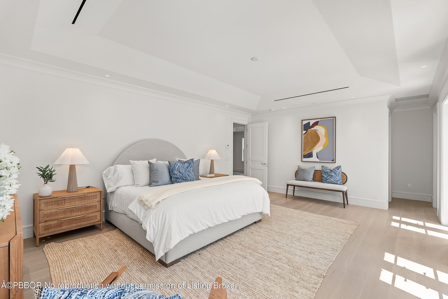 This is a bright and airy primary bedroom featuring a large bed with a light gray upholstered headboard and white bedding accented with blue pillows. The room is decorated in a minimalist style with light wood floors, a jute rug, and a neutral color palette. A bench sits beneath an abstract painting, and natural light floods the space, creating a serene and inviting atmosphere.