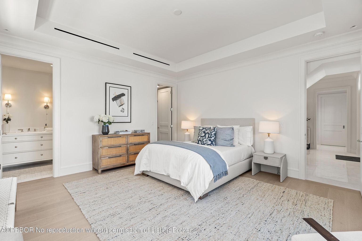 This is a bright and airy primary bedroom featuring a neutral color palette. The room includes a bed with blue accent pillows and a throw, a wooden dresser with decorative items, and a light gray nightstand with a lamp. The space is well-lit and inviting, with a large area rug adding texture to the wooden floors.