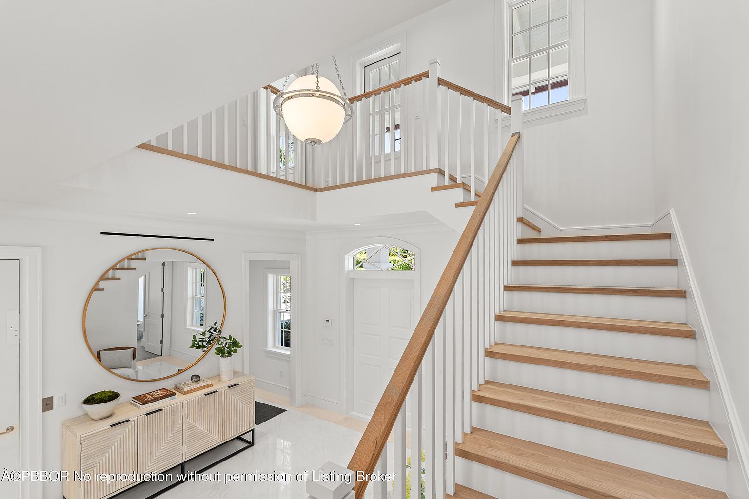 This interior shot showcases a bright and airy hallway with a staircase. The staircase features wooden treads and white risers, complemented by a wooden handrail and white balusters. A modern, spherical light fixture hangs from the ceiling, adding a touch of elegance to the space, while a round mirror and console table decorate the hallway.