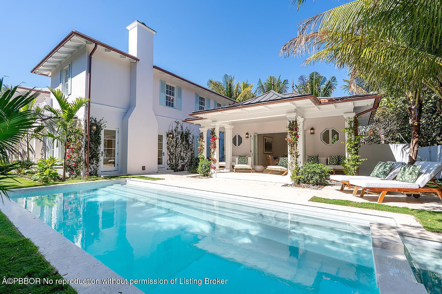 This image showcases a luxurious backyard featuring a pristine pool with crystal-clear water, surrounded by lush greenery and palm trees. The elegant white house with light blue shutters provides a stunning backdrop, complemented by a covered patio area with comfortable seating and sun loungers. The scene exudes a sense of relaxation and upscale outdoor living.