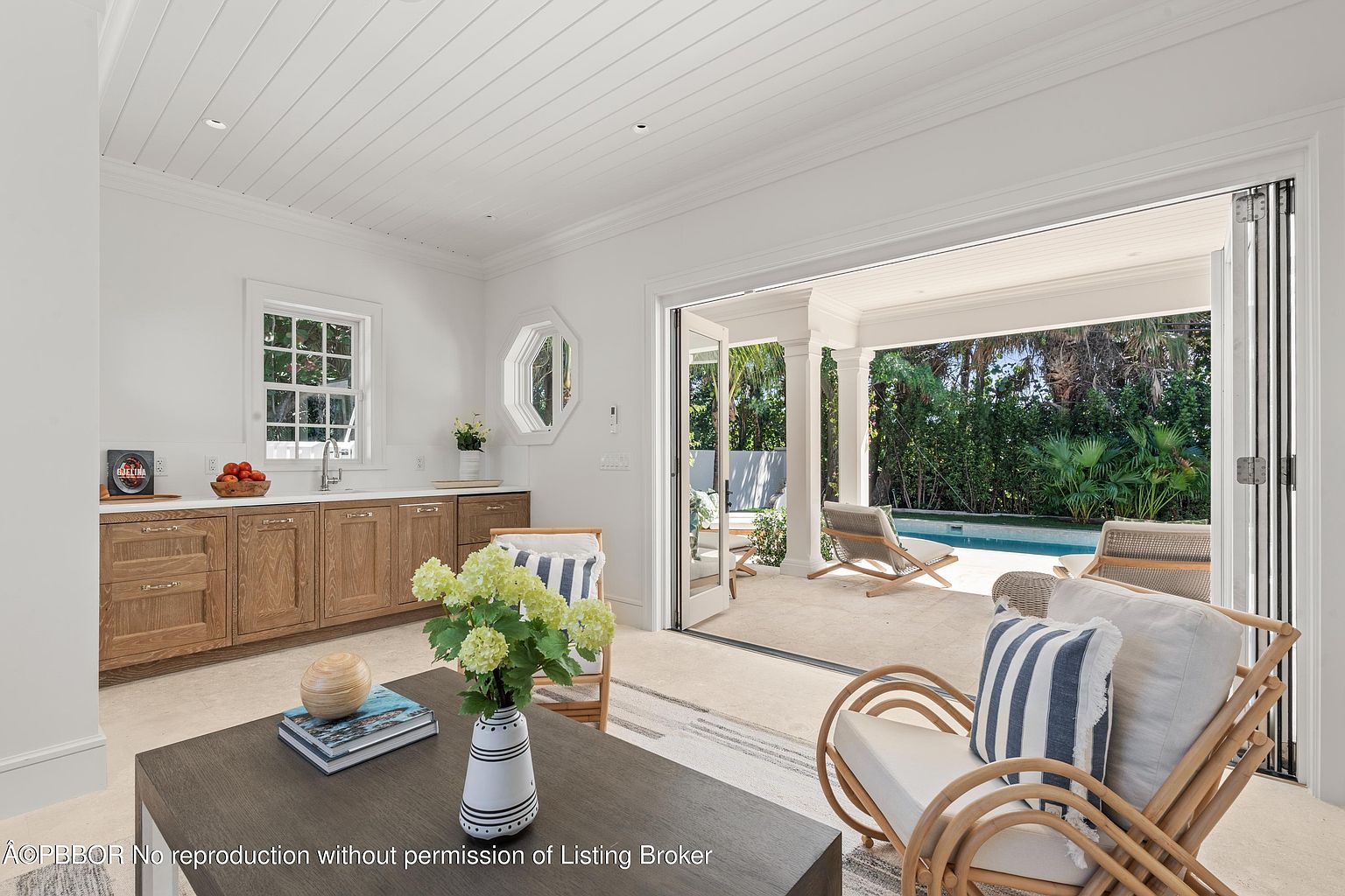 This interior shot showcases a bright and airy living room that seamlessly transitions to an outdoor patio and pool area through large, folding glass doors. The room features built-in wooden cabinetry, comfortable rattan seating with striped cushions, and a stylish coffee table with decorative objects. The design emphasizes indoor-outdoor living, creating a relaxed and inviting atmosphere.