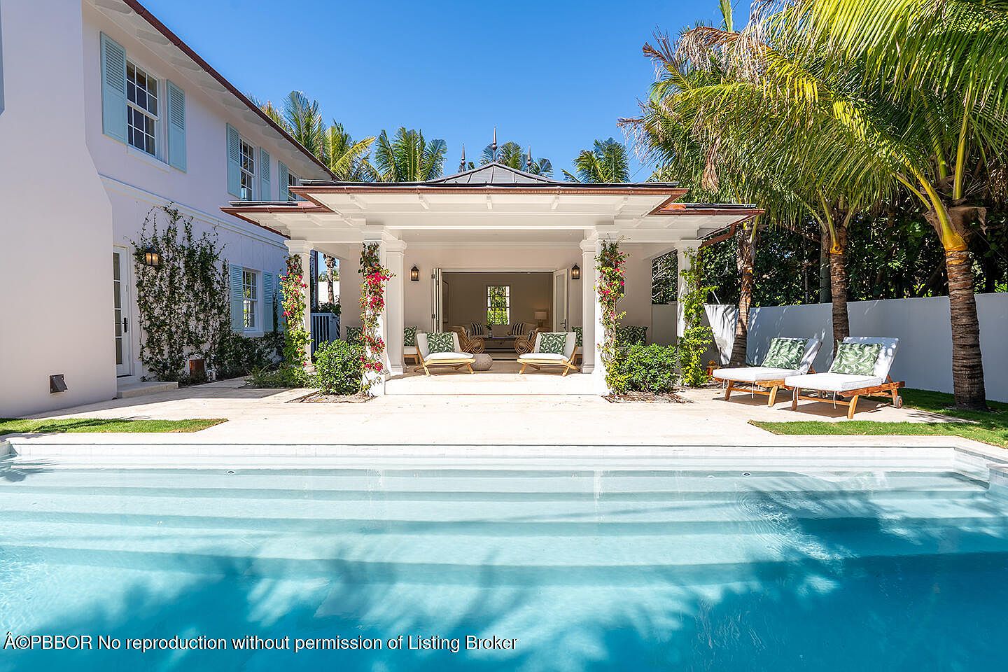 This image showcases a luxurious backyard featuring a pristine pool with clear blue water and steps leading into it. A covered patio area with elegant columns and comfortable seating overlooks the pool, while lush tropical landscaping and palm trees add to the serene atmosphere. The overall impression is one of relaxation and upscale outdoor living.