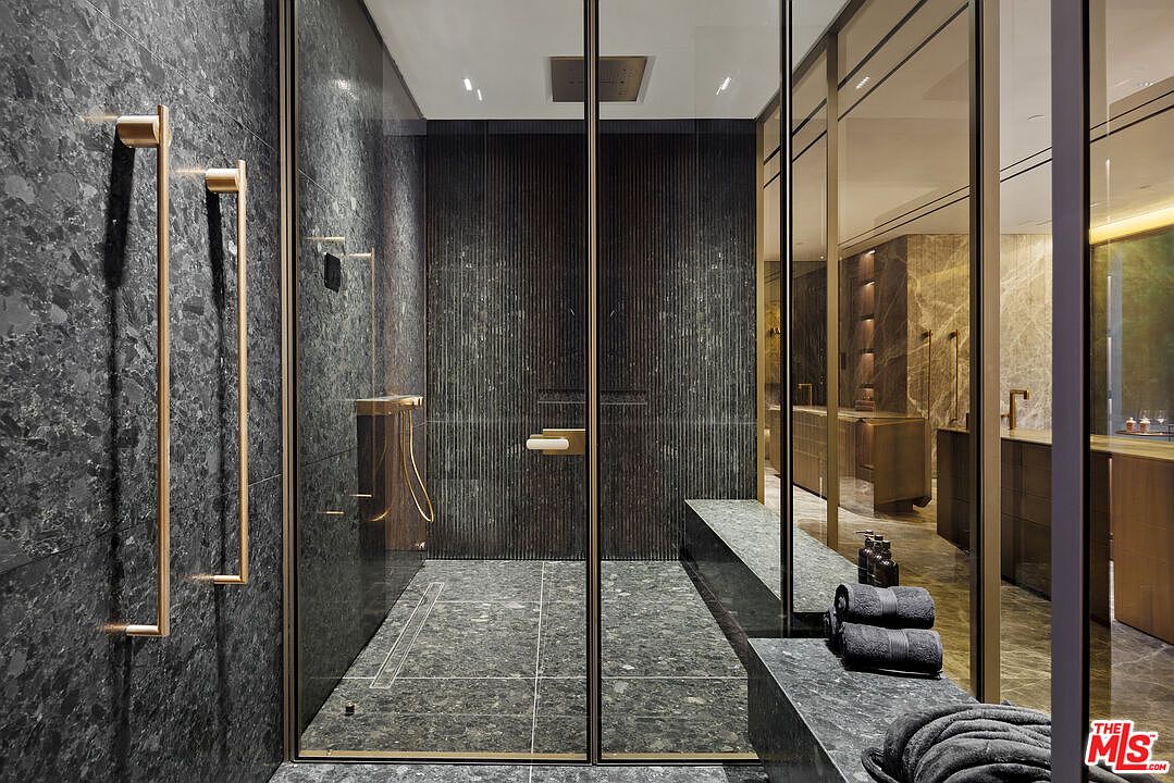 This luxurious primary bathroom features a spacious walk-in shower enclosed by sleek, dark-framed glass panels, showcasing sophisticated dark stone tiling throughout. The design is highlighted by elegant gold-toned fixtures, including a vertical towel rack and shower hardware, which contrast beautifully against the moody, textured stone walls. A built-in stone bench and minimalist aesthetic create a high-end, spa-like atmosphere perfect for a modern luxury residence.