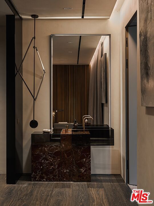 This sophisticated powder room features a striking vanity crafted from dark, veined marble, paired with a sleek, minimalist glass sink and modern brass faucet. A large, rectangular mirror reflects the warm, ambient lighting and elegant drapery, while a unique geometric pendant light adds a sculptural element to the space. The overall aesthetic is one of high-end, moody luxury, emphasizing rich textures and contemporary design.