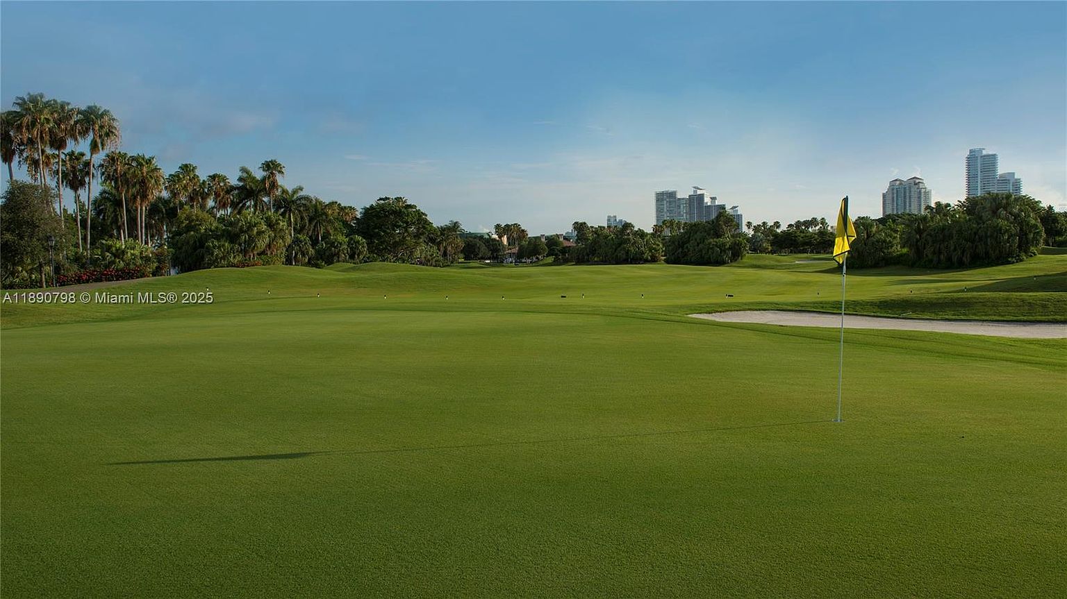 This image showcases a lush green golf course under a clear blue sky, with palm trees lining the perimeter and modern buildings visible in the distance. A yellow flag marks a hole on the green, and a sand trap is visible nearby. The scene evokes a sense of luxury and recreation, highlighting the community's amenities.