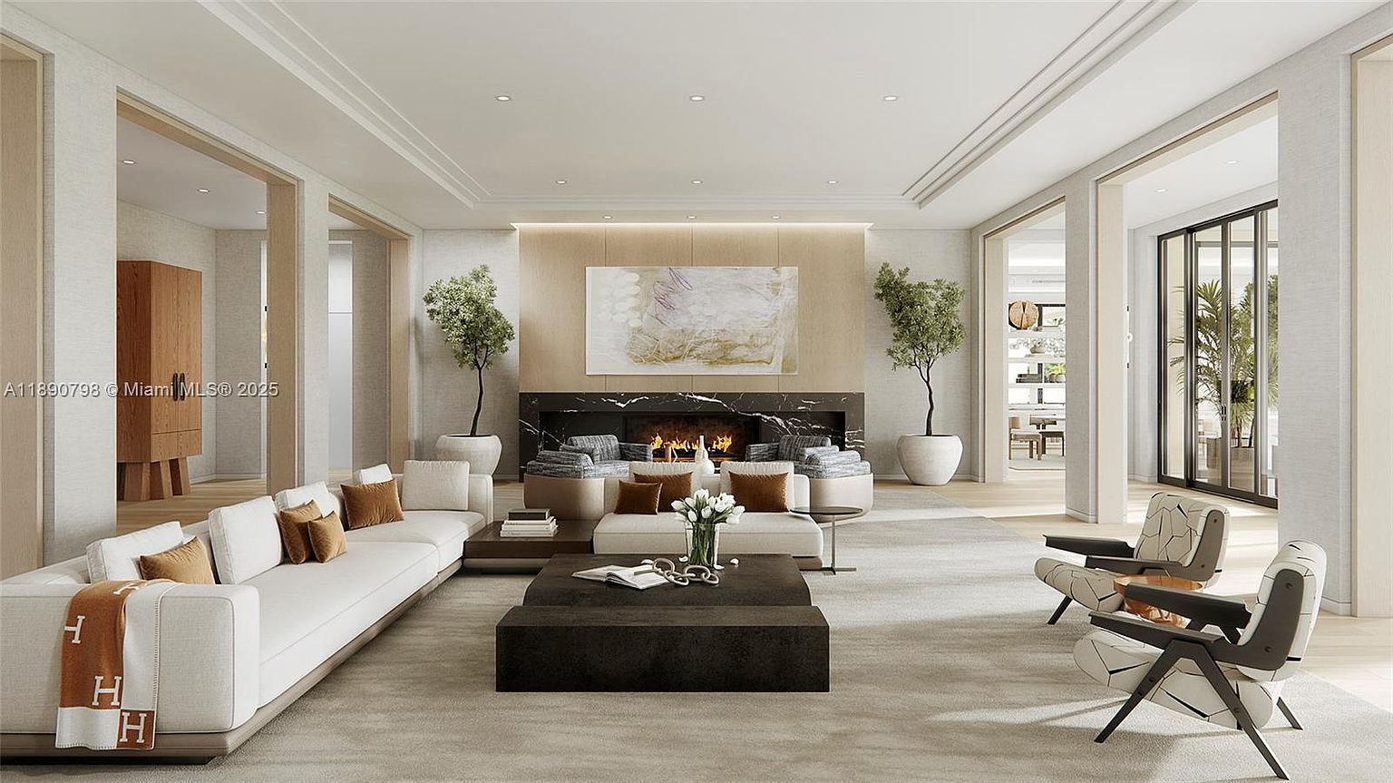 This is a spacious and luxurious living room featuring a long white sofa with decorative pillows and a throw, a dark coffee table, and two modern chairs. A fireplace with a black marble surround is the focal point, flanked by potted trees and artwork. The room is well-lit and exudes a sophisticated and comfortable atmosphere.