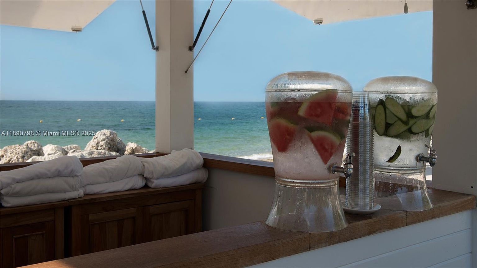 This image showcases a refreshing amenity at a beachside property. Two large beverage dispensers, one with watermelon and the other with cucumber, sit on a wooden counter, with neatly folded white towels nearby. The backdrop features a serene ocean view under a clear blue sky, creating an inviting and luxurious atmosphere.
