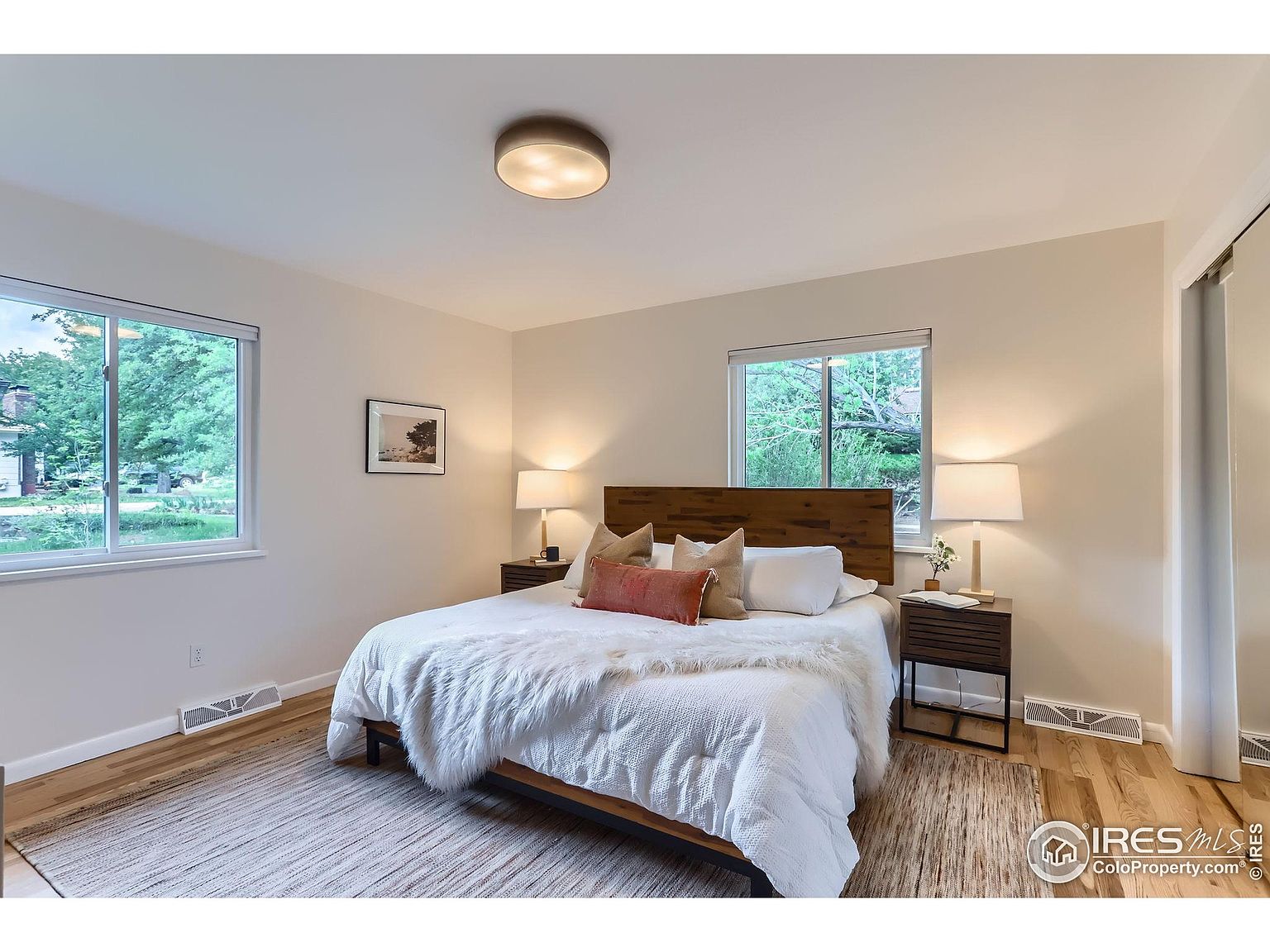 This is a well-lit primary bedroom featuring a wooden headboard bed with white linens and decorative pillows. The room has two windows providing natural light, bedside tables with lamps, and a neutral-toned rug. The overall impression is cozy and inviting, showcasing a comfortable and stylish sleeping space.