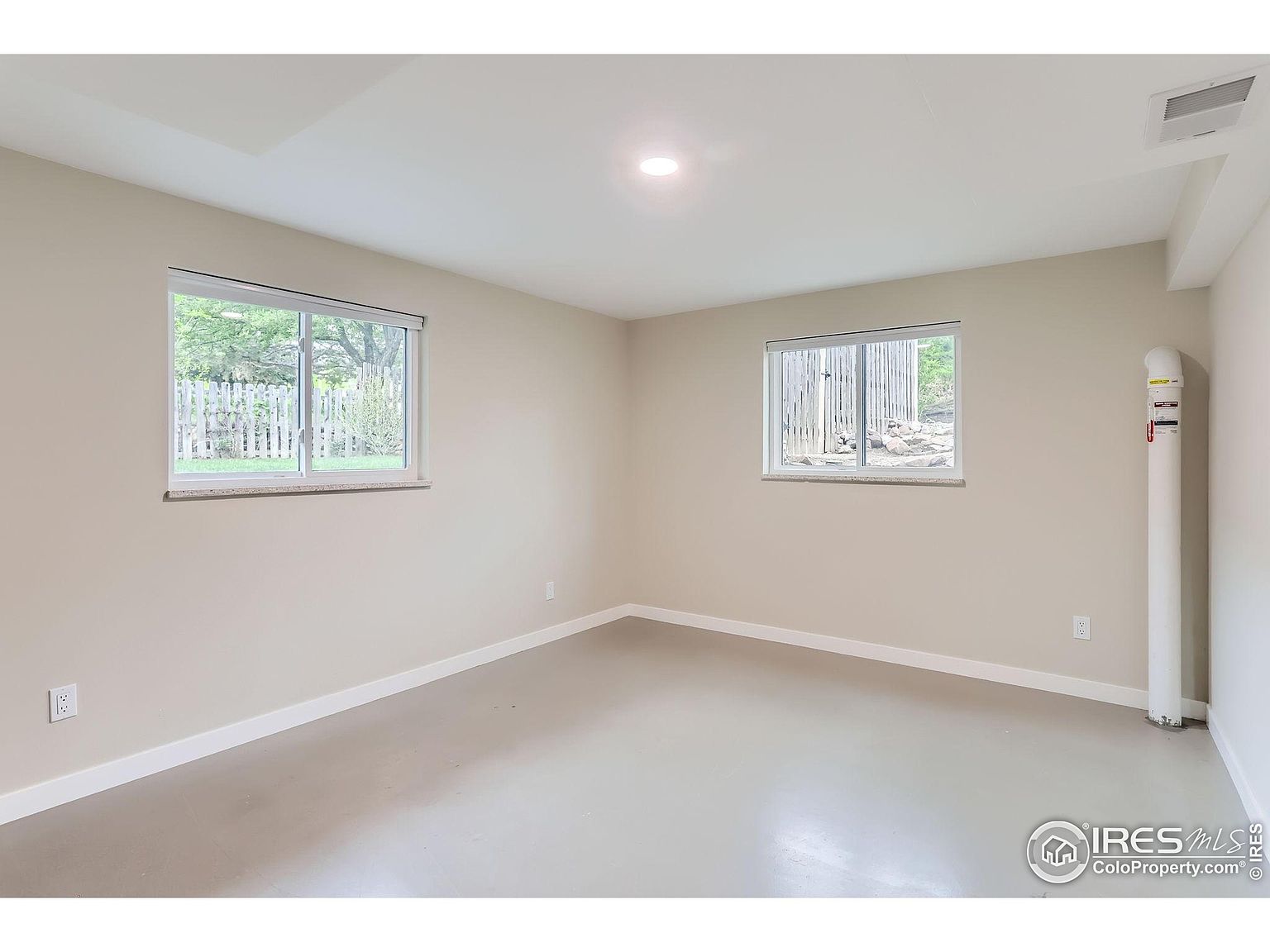 The image showcases a well-lit basement room with neutral-toned walls and a light gray floor. Two windows provide natural light, and a white pipe runs vertically along the right wall. The room appears clean and spacious, offering potential for various uses.