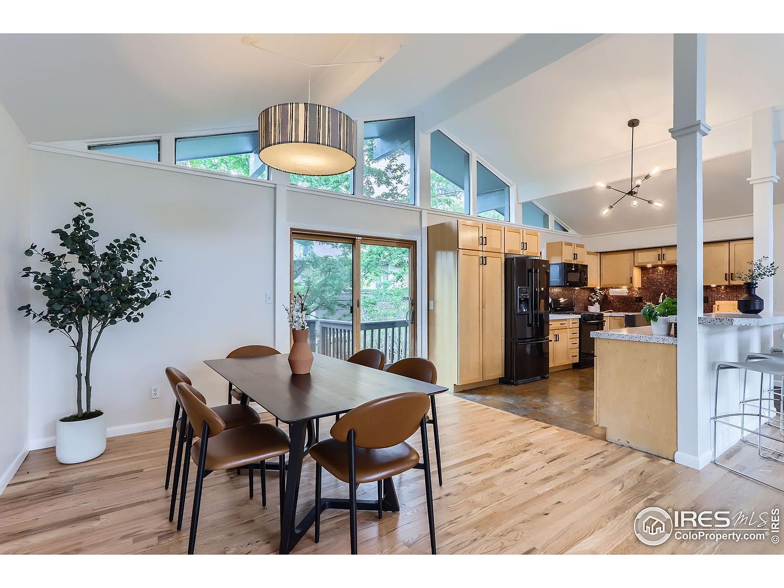 This interior shot showcases a dining area adjacent to a kitchen. The dining space features a modern table with chairs, a decorative tree, and a unique light fixture. The kitchen has light wood cabinets, black appliances, and an open layout, creating a seamless flow between the two spaces.