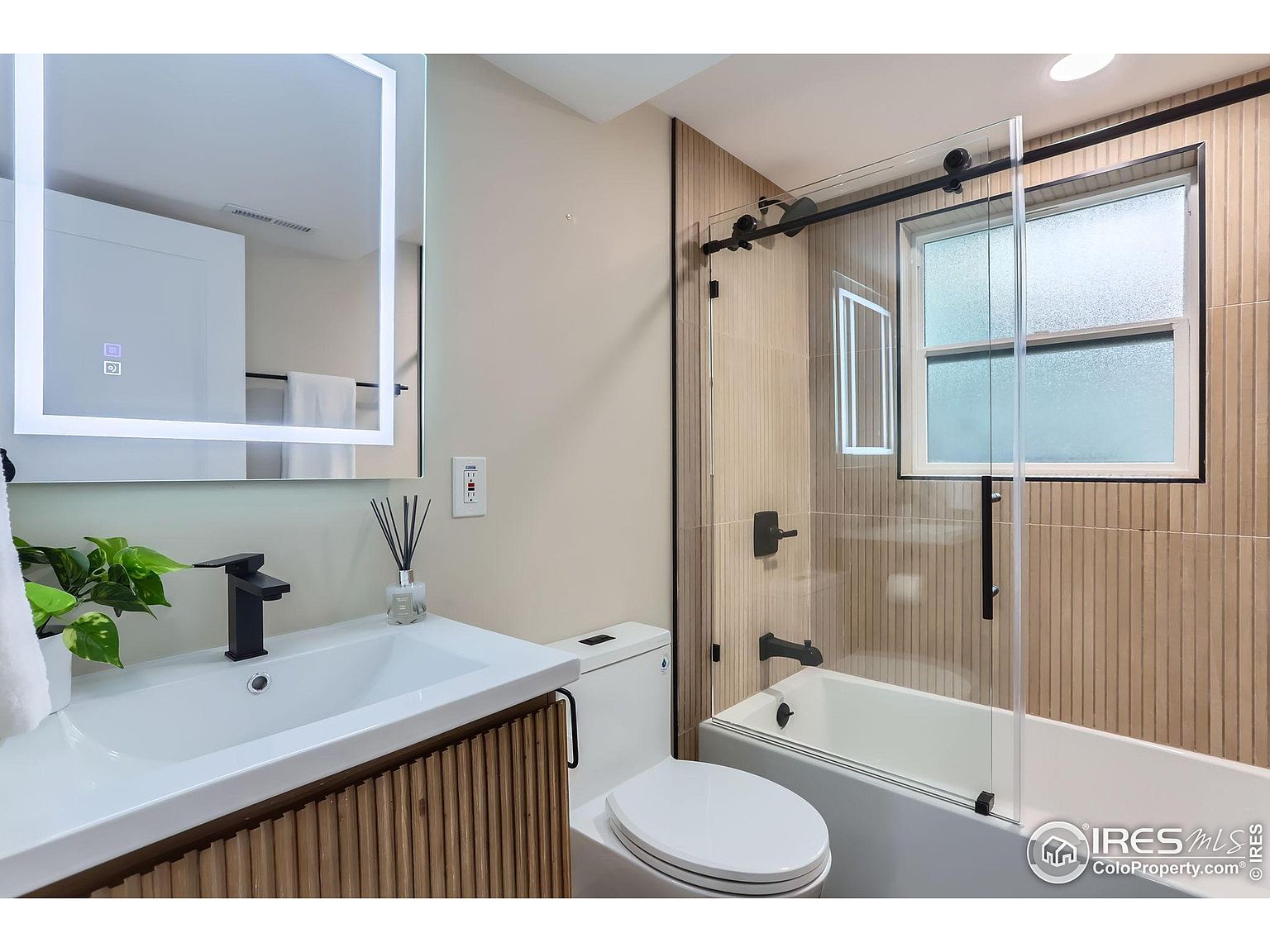 This is a well-lit bathroom featuring a modern design. The vanity has a white countertop and a wooden cabinet with vertical slats. A sleek, black faucet complements the white sink. The bathroom includes a toilet and a bathtub with a glass shower door, framed in black, and a window. The walls are tiled with a light brown, vertically-oriented pattern, adding texture and visual interest.