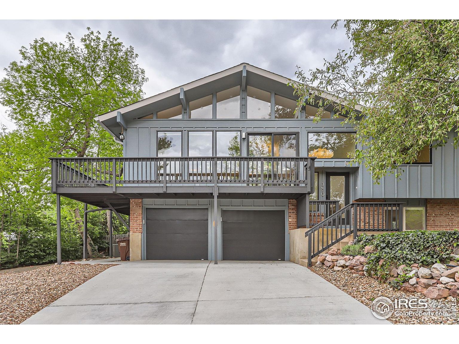 This is a front view of a two-story house with a modern architectural style. The house features a large deck above the garage, accessible from the main level. The exterior is painted in a muted blue-gray tone, complemented by dark gray garage doors and trim. The front yard is landscaped with rocks and greenery, adding to the property's curb appeal.