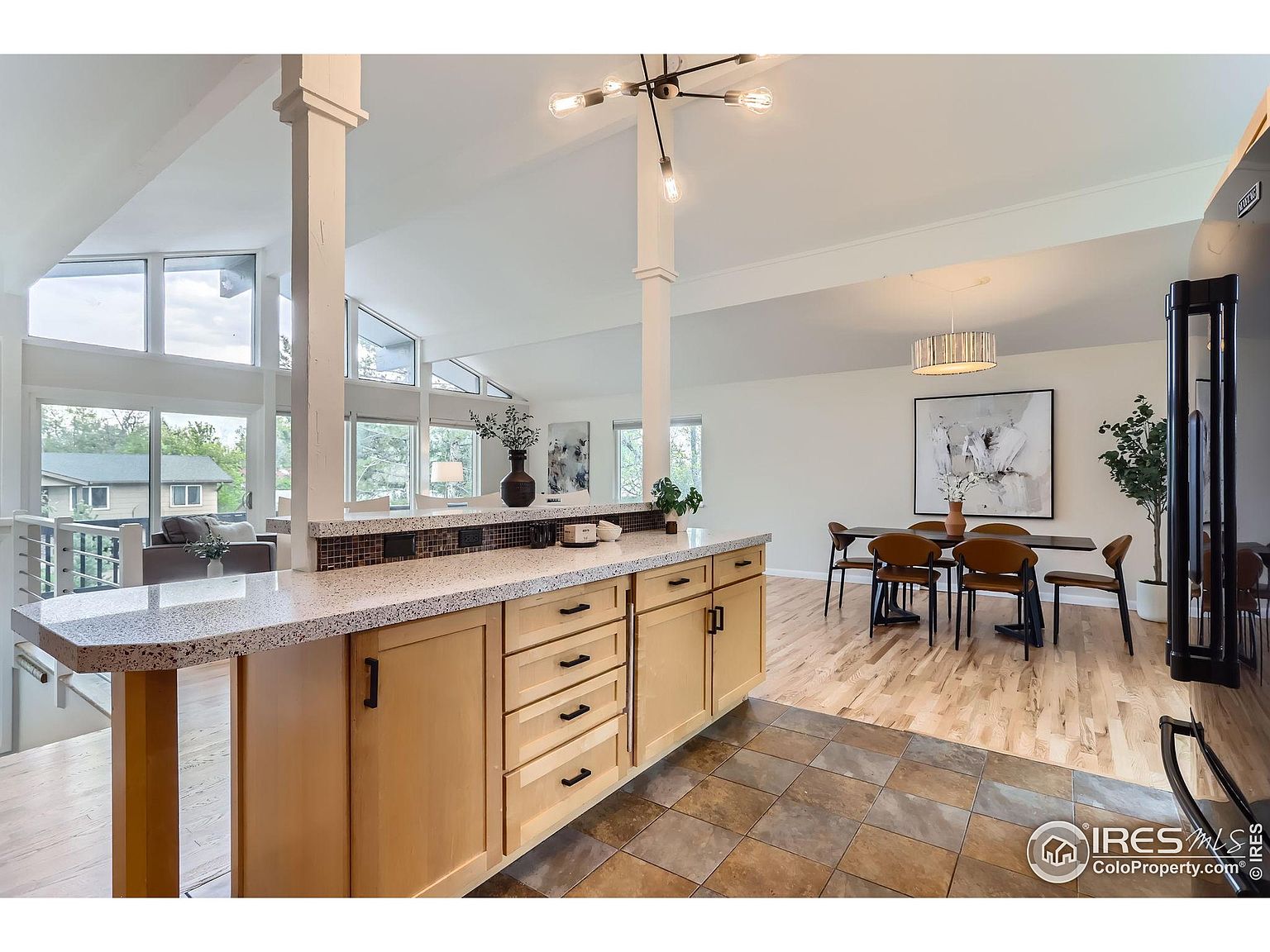 This interior shot showcases a bright and open kitchen and dining area. The kitchen features light wood cabinetry, speckled countertops, and a tiled backsplash, while the dining area includes a modern table set and a large abstract art piece. The space is well-lit with natural light from large windows and modern light fixtures, creating a welcoming and airy atmosphere.