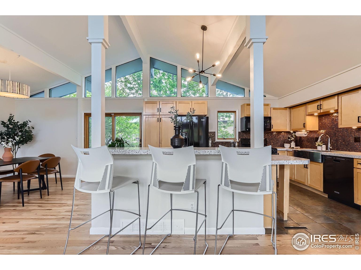 This is an interior shot of a modern kitchen with a high, vaulted ceiling featuring large windows that let in natural light. The kitchen includes a breakfast bar with three white bar stools, light wood cabinetry, black appliances, and a dark backsplash. The flooring transitions from light wood in the foreground to tile in the kitchen area, creating a warm and inviting space.