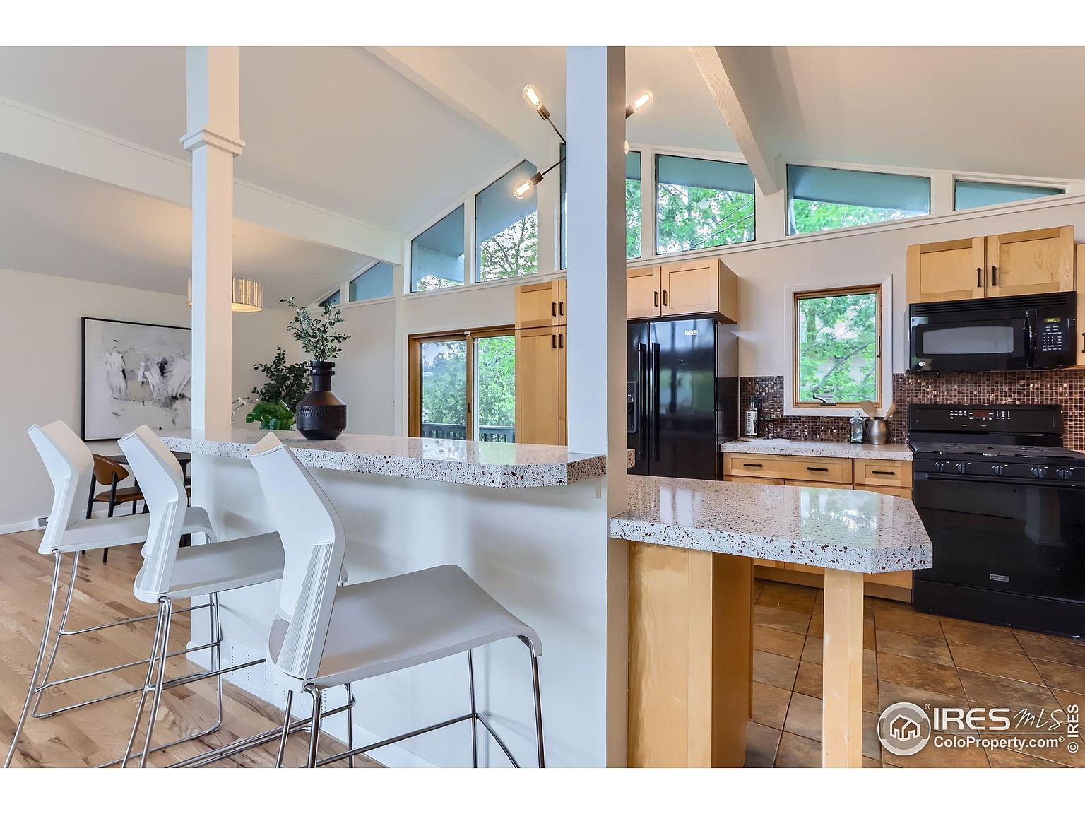 This is an interior shot of a kitchen featuring light wood cabinets, black appliances, and a speckled countertop that extends into a breakfast bar area with white bar stools. The kitchen has a modern, open feel with clerestory windows providing natural light and a view to the outside. The flooring is a mix of wood and tile, adding visual interest.