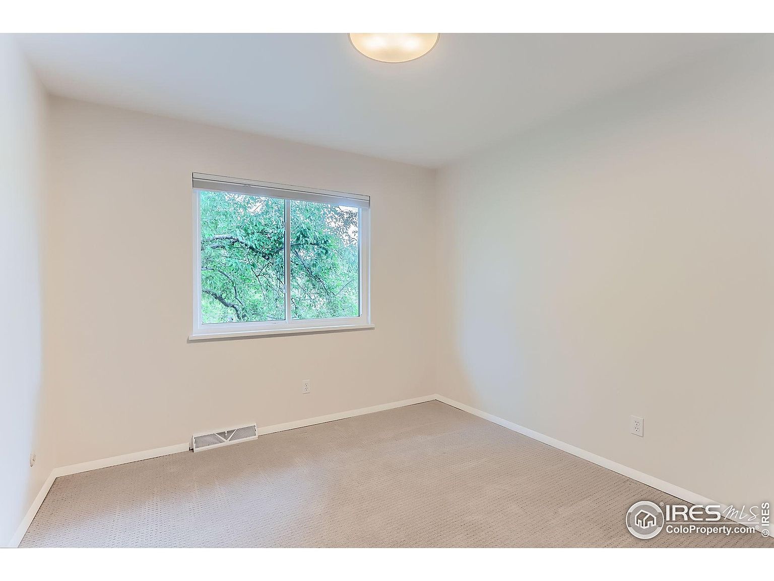The image showcases a bright and airy bedroom with neutral-toned walls and carpet. A window provides natural light and a view of greenery outside. The room appears clean and well-maintained, offering a blank canvas for potential buyers to envision their own style.