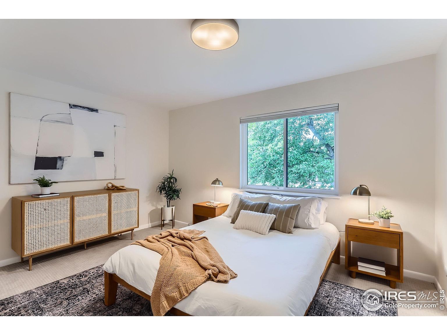 This is a bright and inviting primary bedroom featuring a neutral color palette. The room includes a bed with white linens and decorative pillows, two wooden nightstands with lamps, and a wooden cabinet with woven doors. A large abstract painting hangs above the cabinet, and a window provides natural light and a view of greenery outside.