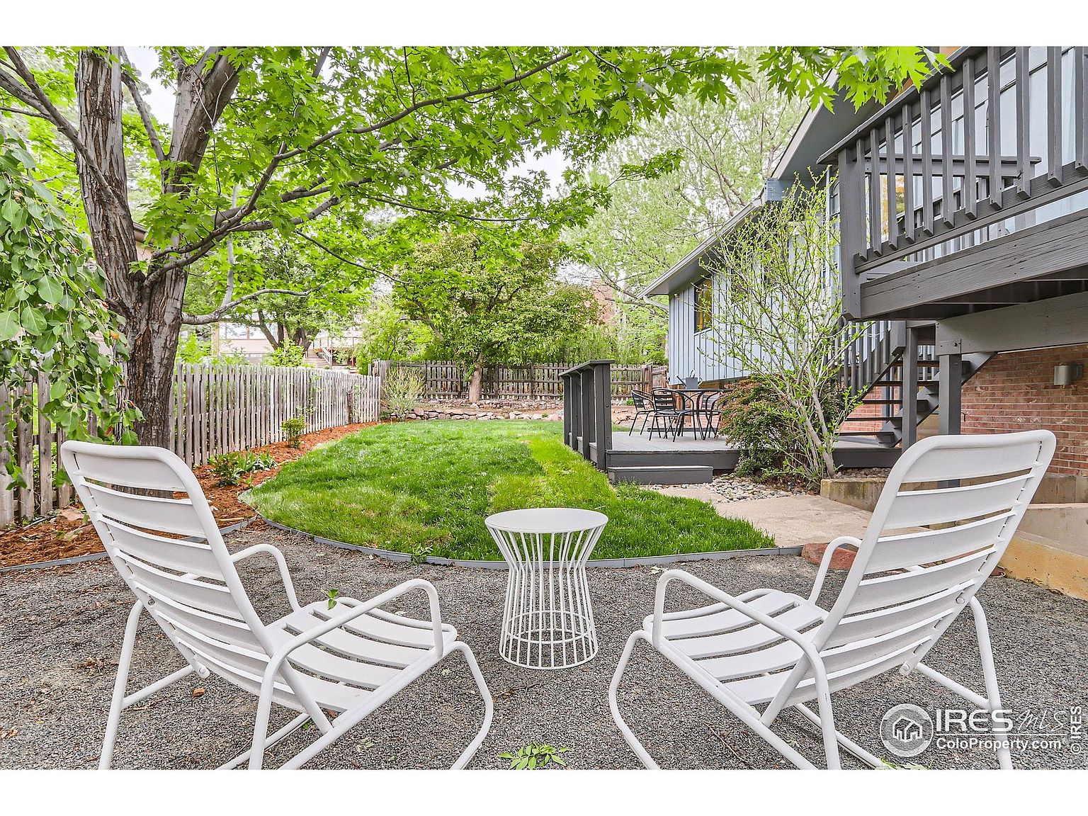 This is a serene backyard featuring a well-maintained lawn, mature trees providing shade, and a cozy seating area with white chairs and a small table. A wooden fence encloses the yard, offering privacy, while a deck extends from the house, creating an inviting outdoor living space. The overall impression is one of tranquility and relaxation.