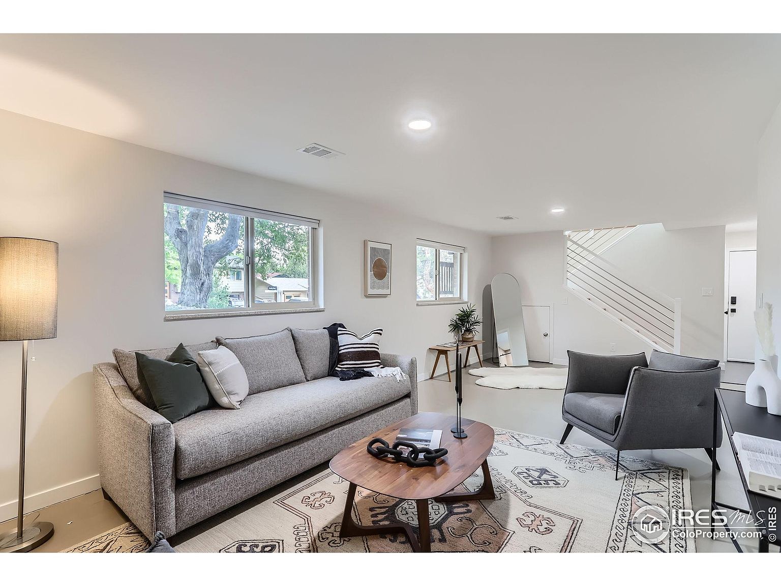 This is a well-lit living room featuring a gray sofa with decorative pillows, a unique wooden coffee table, and a gray armchair. The room is decorated with a patterned area rug, a floor lamp, and various decorative items. A staircase is visible in the background, contributing to the modern and inviting atmosphere of the space.