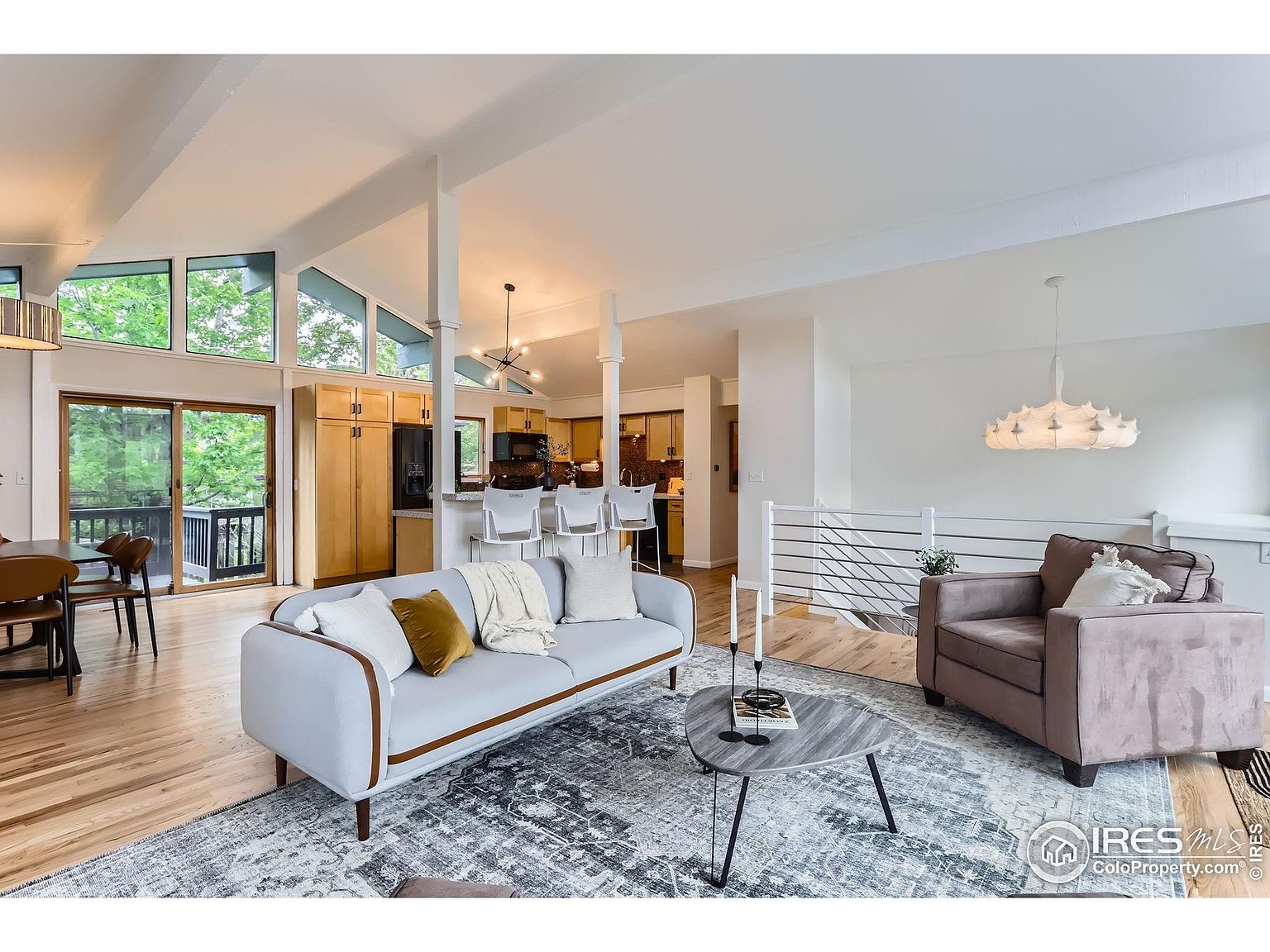 This is an interior shot of a living room featuring a light gray sofa with decorative pillows, a modern coffee table, and a brown armchair. The room is well-lit with natural light coming through large windows and a sliding glass door, and the hardwood floors and neutral color palette create a bright and airy atmosphere. The open floor plan connects the living room to the kitchen area, enhancing the sense of spaciousness.