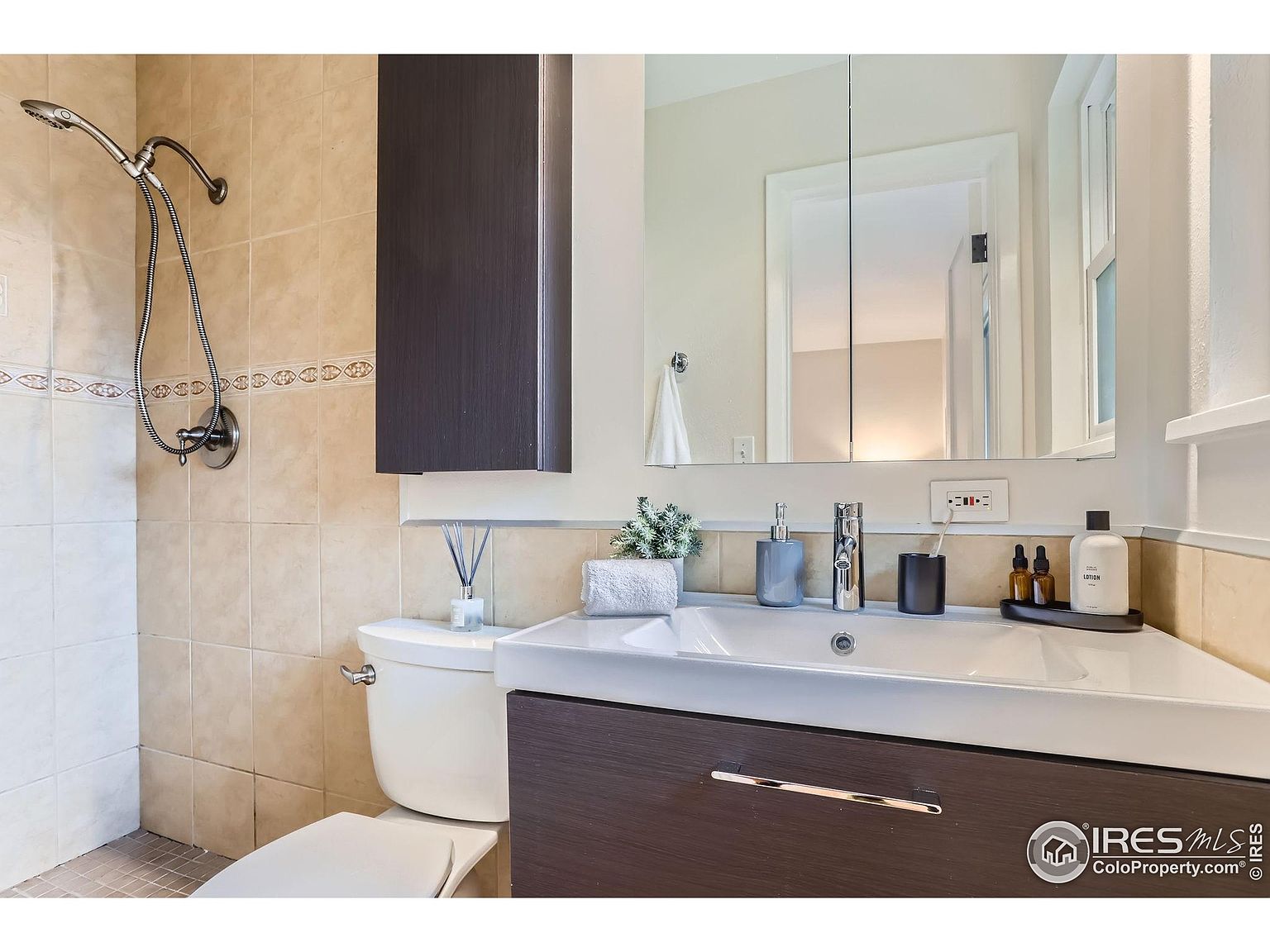 This is a well-lit bathroom featuring a modern vanity with a dark wood finish and a white countertop with an integrated sink. A mirrored medicine cabinet hangs above the vanity, reflecting the light and creating a sense of spaciousness. The shower area is tiled in a neutral tone, and a handheld showerhead is visible.