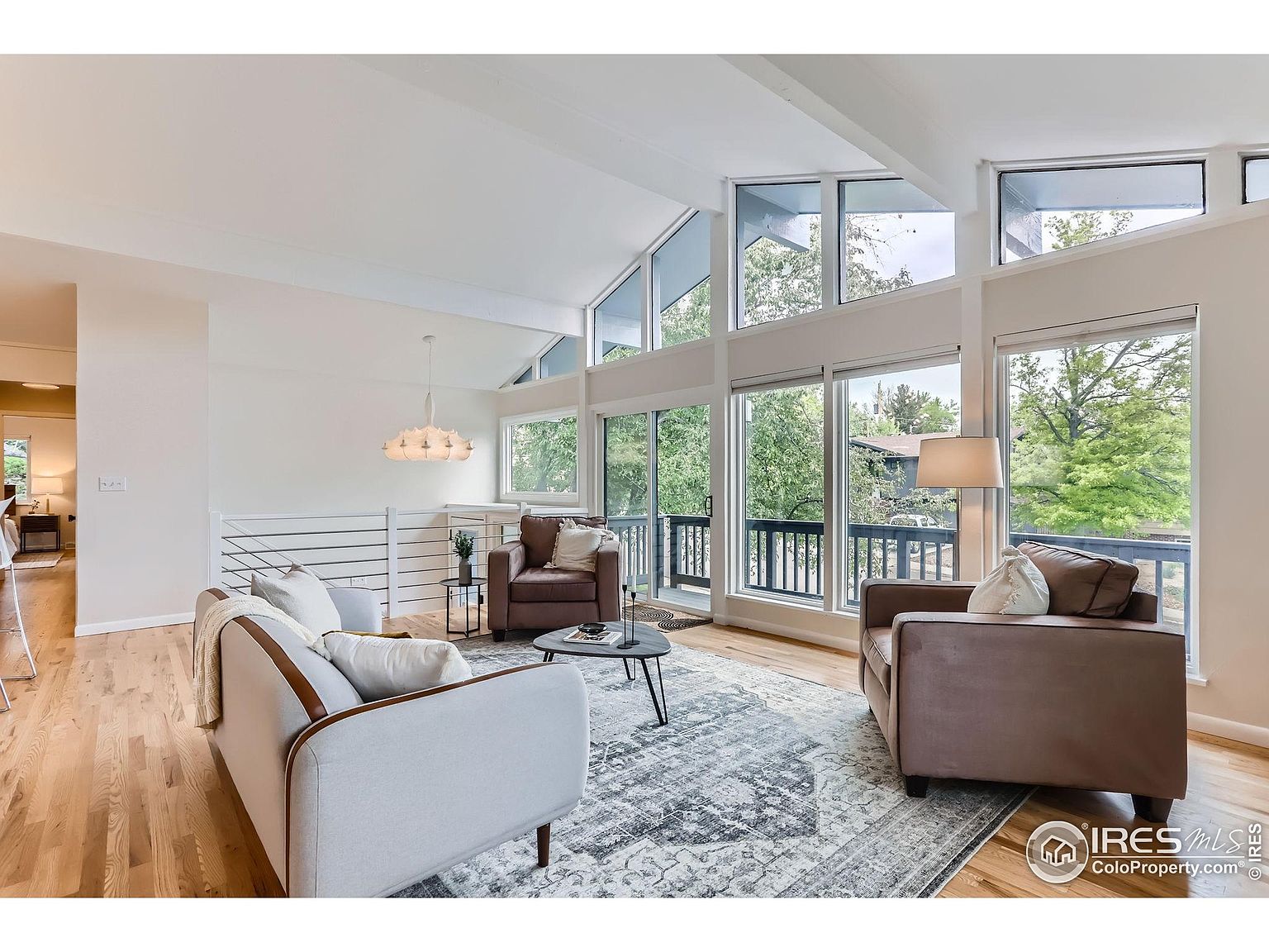 This is an interior shot of a living room featuring a modern design with large windows that offer ample natural light and views of the outdoors. The room is furnished with a light-colored sofa, two brown armchairs, and a patterned area rug, creating a comfortable and inviting space. The high ceilings and unique window design add architectural interest, making it a desirable living area.