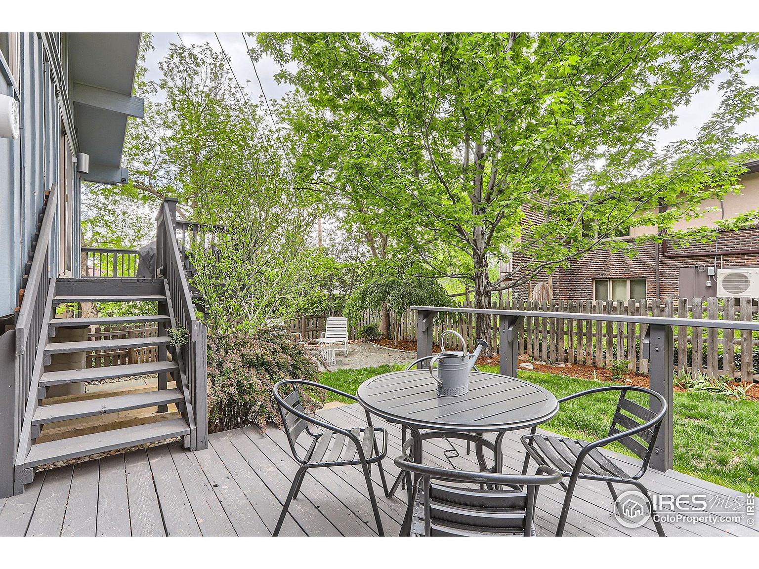 This image showcases a charming outdoor patio area, featuring a gray wooden deck with a round table and four matching chairs. The deck is accessible via a set of stairs and overlooks a well-maintained lawn and garden. A wooden fence and mature trees provide privacy and a sense of tranquility, making it an ideal space for relaxation and outdoor dining.
