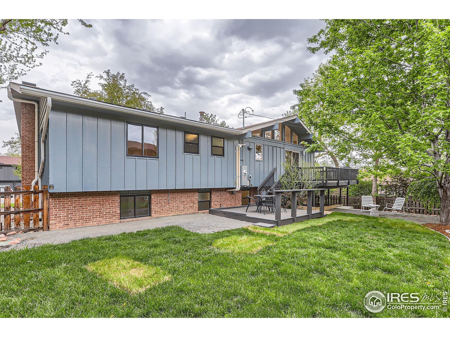 This is a rear view of a two-story house with a combination of brick and blue siding. A wooden deck extends from the upper level, providing an outdoor living space. The backyard features a well-maintained lawn with some patchy areas and a wooden fence, creating a private and inviting atmosphere.