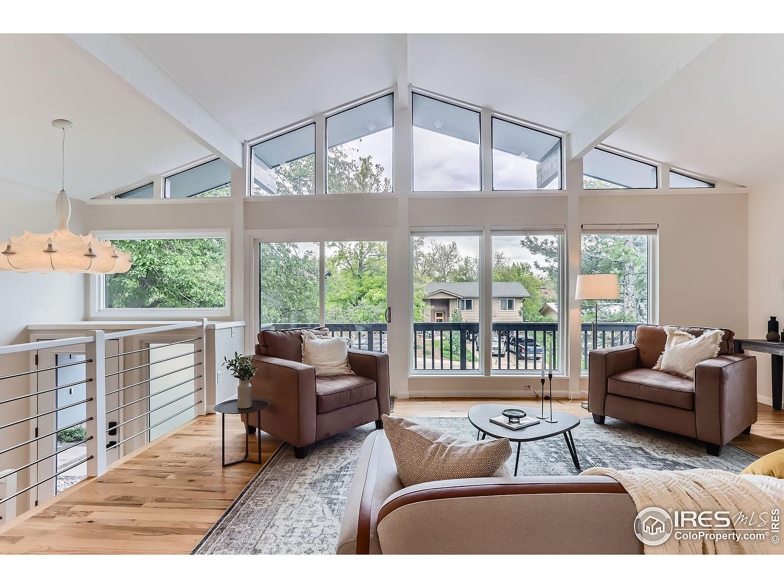 This is an interior shot of a living room featuring a high ceiling with large windows that offer natural light and views of the outdoors. The room is furnished with two brown armchairs, a sofa, and a coffee table, all arranged on a patterned area rug. The space has a modern and comfortable feel, suitable for relaxation and entertaining.