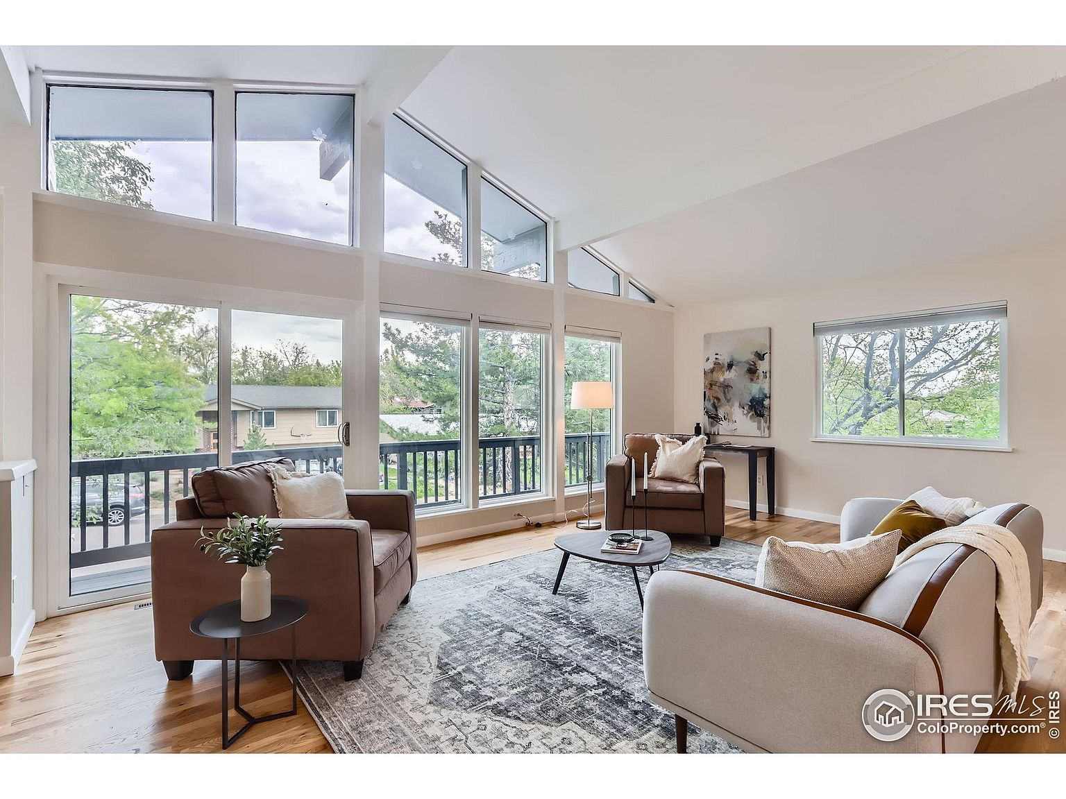 This is a bright and airy living room featuring large windows that offer a view of the outdoors. The room is furnished with comfortable seating, including a brown armchair and a light-colored sofa, arranged around a central rug and coffee table. The high ceilings and neutral color palette create a spacious and inviting atmosphere, perfect for relaxation and entertaining.
