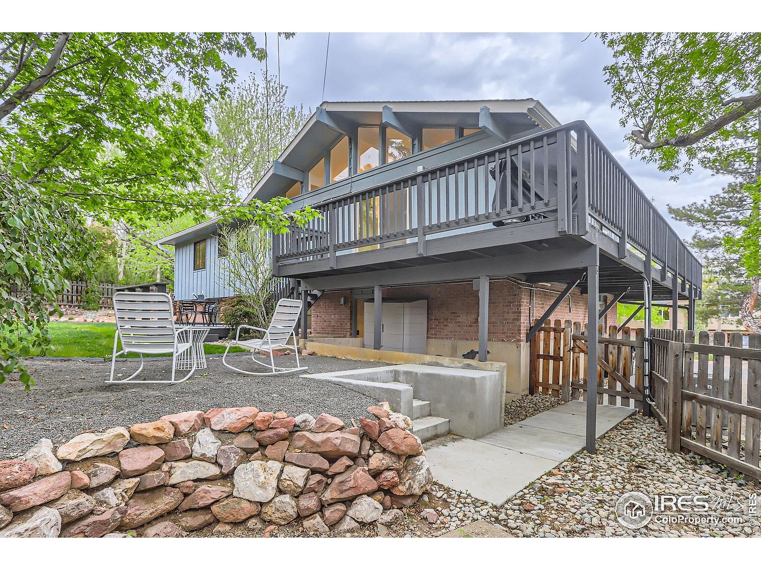 This image showcases the exterior of a two-story home with a prominent deck. The yard features a rock wall, gravel landscaping, and a small patio area with outdoor seating. The overall impression is a well-maintained outdoor space with a blend of natural and architectural elements.