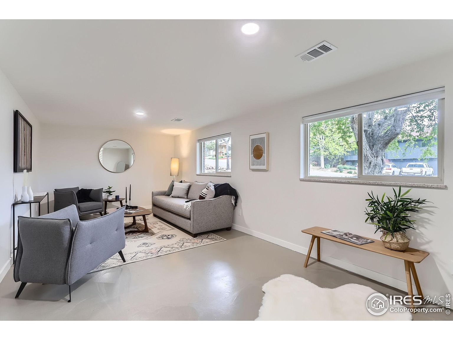 This is an interior shot of a living room featuring a gray sofa and two gray armchairs arranged around a coffee table on a patterned rug. Natural light streams in through two windows, illuminating the light-colored walls and flooring. The room is decorated with modern art and a round mirror, creating a clean and inviting atmosphere.