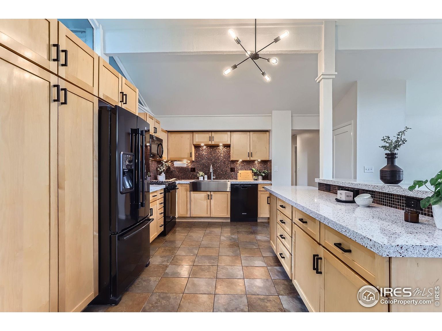 This is a well-lit kitchen featuring light wood cabinetry with black hardware and black appliances. The countertops are light-colored with a speckled pattern, and the backsplash is a mosaic tile. The flooring is a neutral-toned tile, and a modern chandelier hangs from the ceiling.
